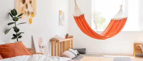 A stylish, modern bedroom in a rental apartment featuring an orange hammock, a wooden bed with rust-colored bedding, and minimalist decor.