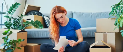 Young woman appearing worried about renting mistakes while she is looking through documents sitting on the floor among moving boxes.