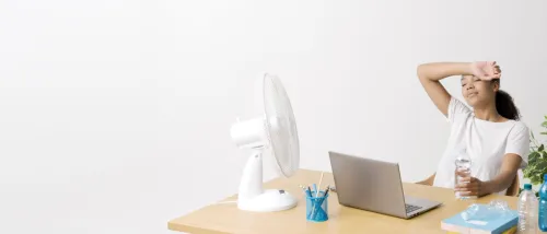 A young renter works from home during a heatwave, looking exhausted as she wipes her forehead, with a fan blowing on her and several water bottles on her cluttered desk.