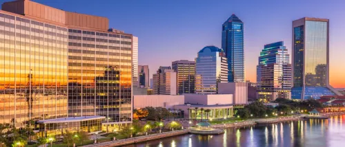 Downtown Jacksonville skyline at sunset, featuring high-rise buildings reflecting the colorful sky and waterfront views.