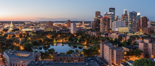 Minneapolis skyline at sunset with tall buildings, lights, and the waterfront.