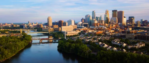 Aerial view of downtown Minneapolis, MN, the most sought-after city for renters in the first half of 2024, featuring a mix of modern high-rise buildings, bridges over the river, and residential areas surrounded by lush greenery under a cloudy sky.