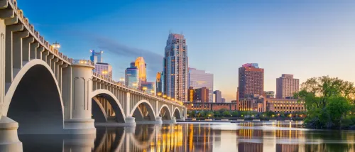 Minneapolis skyline with a bridge at sunset, reflecting in the calm water below.