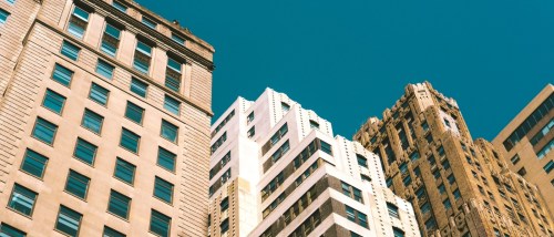 Tall vintage buildings reaching up towards a clear blue sky on a sunny day in New York City, showcasing the contrast of architectural styles and the warmth of sunlight on their facades.