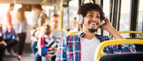 A young man smiles while wearing headphones on a city bus, surrounded by other passengers in a brightly lit interior.