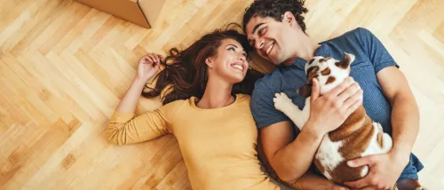 A happy couple lying on the floor with their dog with moving boxes in the background.