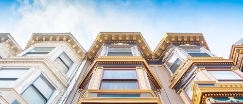 A wide-angle view of the upper facades of three Victorian homes in San Francisco, featuring intricate trim, bright colors, and ornate decorative elements under a clear sky.